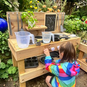 May include: A wooden play kitchen with a chalkboard sign that says "Treaty 3&cent;". The kitchen has a sink, stovetop, and shelves. A child is playing with the kitchen.