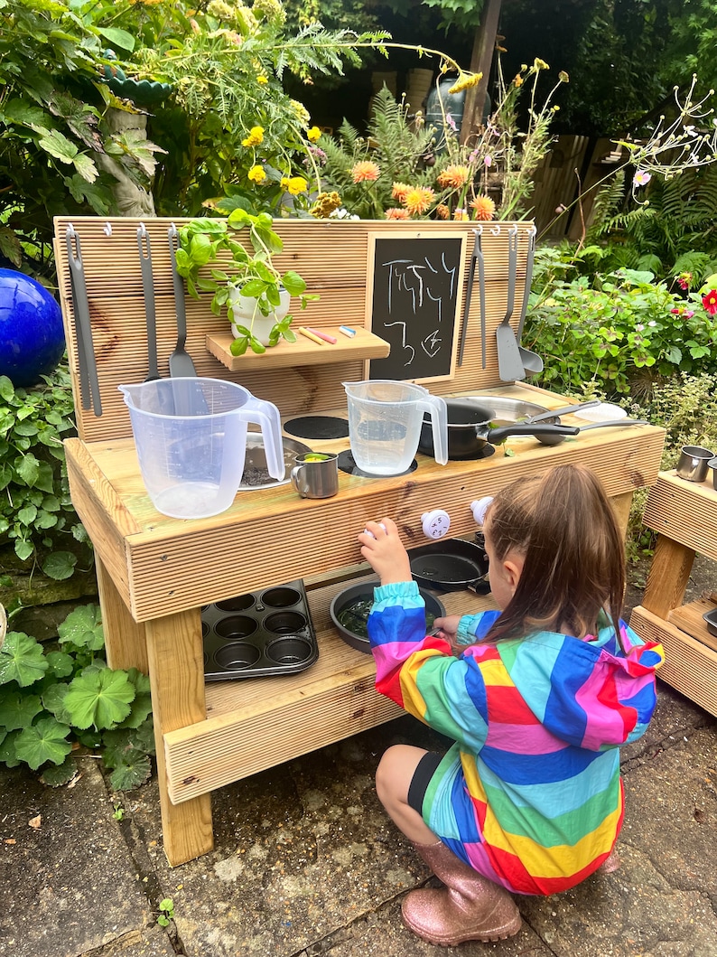 May include: A wooden play kitchen with a chalkboard sign that says "Treaty 3&cent;". The kitchen has a sink, stovetop, and shelves. A child is playing with the kitchen.