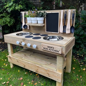 May include: Wooden outdoor play kitchen with a chalkboard, two stainless steel sinks, and a shelf for storage. The kitchen has a sign that reads "Mia's Mud Kitchen".