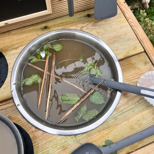 May include: A stainless steel bowl filled with water, green leaves, and wooden sticks. The bowl is sitting on a wooden surface.