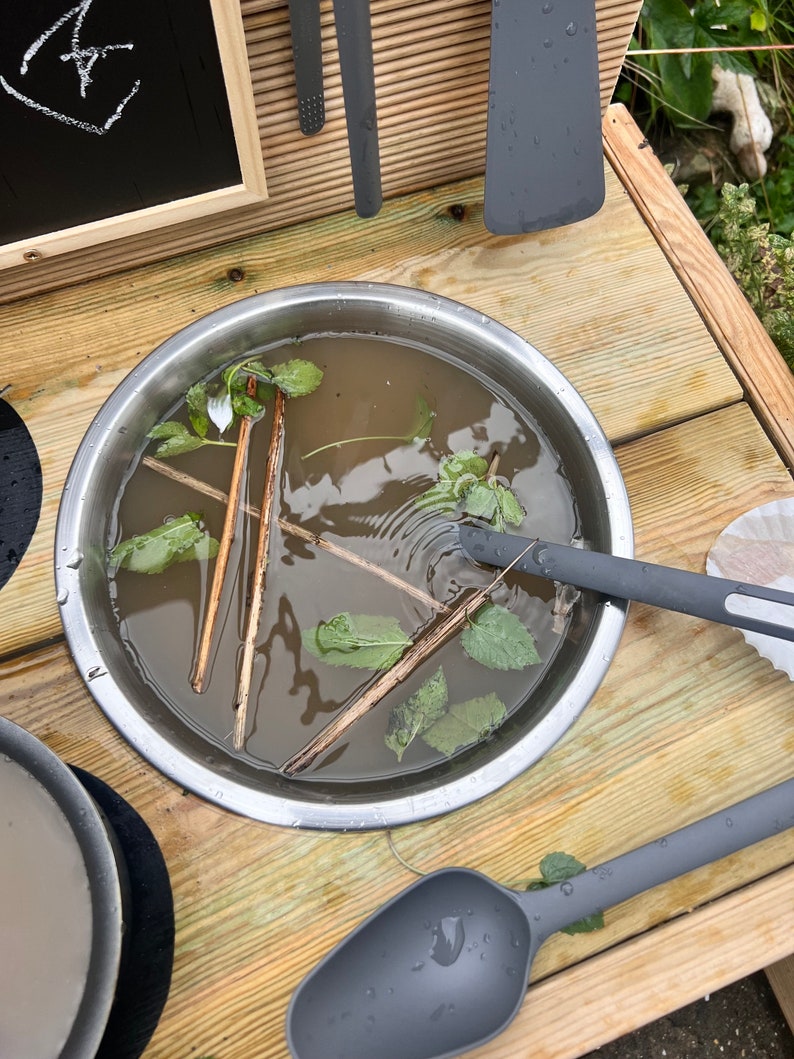 May include: A stainless steel bowl filled with water, green leaves, and wooden sticks. The bowl is sitting on a wooden surface.