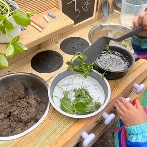 May include: A wooden play kitchen with a black chalkboard, a stainless steel sink, and a set of four black stovetop burners. A child is playing with a spatula and a bowl of green leafy vegetables.