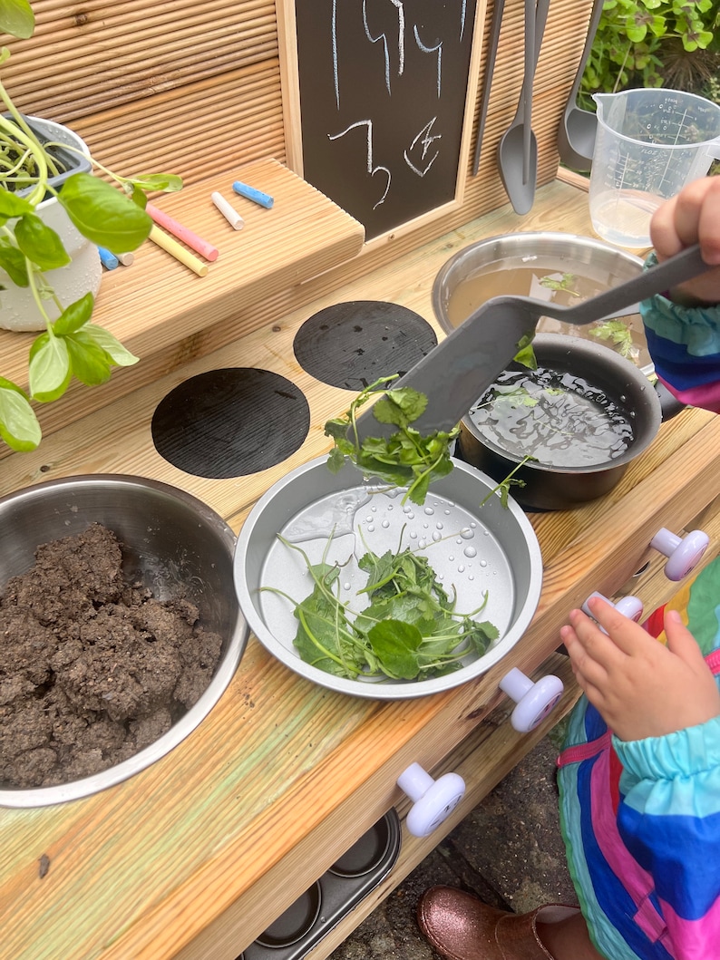 May include: A wooden play kitchen with a black chalkboard, a stainless steel sink, and a set of four black stovetop burners. A child is playing with a spatula and a bowl of green leafy vegetables.