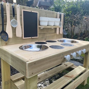May include: Wooden outdoor play kitchen with a black chalkboard, a stainless steel sink, and four white knobs. The kitchen has a shelf with three white bowls and a variety of wooden and metal utensils hanging on hooks.