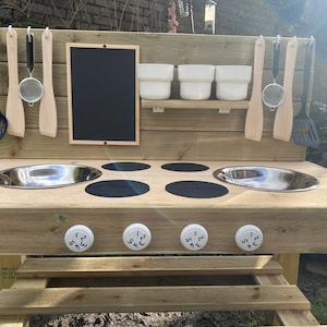 May include: Wooden outdoor play kitchen with two stainless steel sinks, four black stovetop burners, and four white knobs with numbers 1-2-3-4. The kitchen has a chalkboard, a shelf with three white bowls, and hooks for hanging utensils.