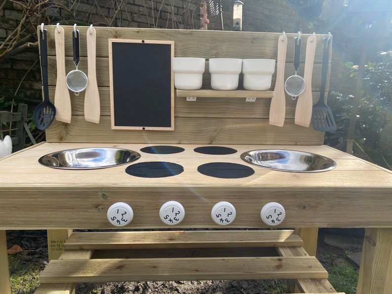 May include: Wooden outdoor play kitchen with two stainless steel sinks, four black stovetop burners, and four white knobs with numbers 1-2-3-4. The kitchen has a chalkboard, a shelf with three white bowls, and hooks for hanging utensils.