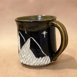May include: Black ceramic mug with a mountain and star design. The mug features a carved white mountain range against a black background. The handle and rim have a brown glaze. A star and a stylized snowflake are also visible.