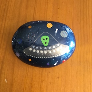 May include: A hand-painted rock depicting a UFO with a green alien face in the center. The rock is painted to look like space with stars, planets, and a comet.