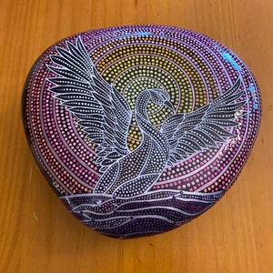 Pode incluir: Uma pedra pintada à mão com um padrão de pontos pretos e brancos que representa um pássaro com as asas abertas. O fundo é um pôr do sol colorido com tons de rosa, roxo, amarelo e laranja.