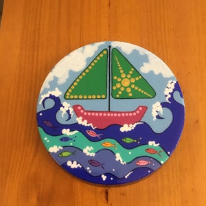 Pode incluir: Uma pintura redonda de madeira de um veleiro verde com casco rosa navegando em águas azuis com ondas brancas e peixes coloridos. O céu é azul com nuvens brancas.