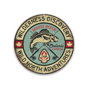 May include: A round, rustic-looking metal sign with a vintage design. It features a stylized illustration of a fish with crossed fishing rods, a camp logo, and the text "WILDERNESS DISCOVERY MOOSE RIVER Ontario CANADA WILD NORTH ADVENTURES".
