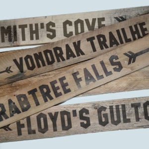 May include: Four wooden signs with black painted arrows and text. The signs read "Smith's Cove Trailhead", "Vondrak Trailhead", "Crabtree Falls", and "Floyd's Gulch".