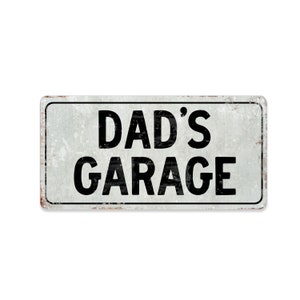 May include: A white metal sign with a distressed finish. The sign reads "DAD'S GARAGE" in black letters.