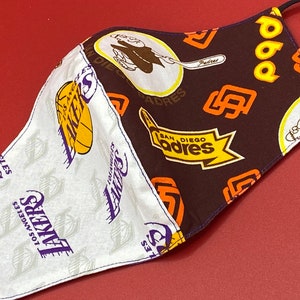Puede incluir: Una mascarilla facial de tela blanca y marrón con ribete morado. Un lado presenta un patrón repetido del logotipo de los Los Angeles Lakers. El otro lado presenta un patrón repetido del logotipo de los San Diego Padres.