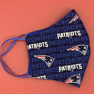 Puede incluir: Una mascarilla facial azul con un patr&oacute;n repetido del logotipo de los New England Patriots y la palabra "Patriots". La mascarilla tiene lazos para las orejas ajustables.