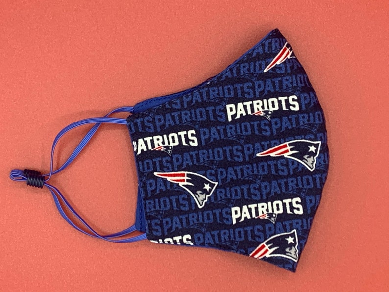 Puede incluir: Una mascarilla facial azul con un patr&oacute;n repetido del logotipo de los New England Patriots y la palabra "Patriots". La mascarilla tiene lazos para las orejas ajustables.