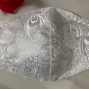 Puede incluir: Máscara facial blanca con un patrón floral blanco. La máscara está hecha de una tela suave y sedosa.