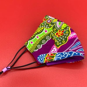Puede incluir: Una mascarilla facial de color morado, verde y rosa con lazos elásticos negros para las orejas y ribete morado. La mascarilla tiene un diseño floral con un patrón paisley.