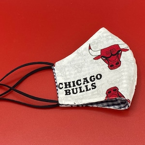 Puede incluir: M&aacute;scara facial blanca con un patr&oacute;n a cuadros blanco y negro en el interior. El exterior de la m&aacute;scara presenta un fondo blanco con un logotipo rojo de los Chicago Bulls y el texto "CHICAGO BULLS". La m&aacute;scara tiene lazos el&aacute;sticos negros para las orejas.
