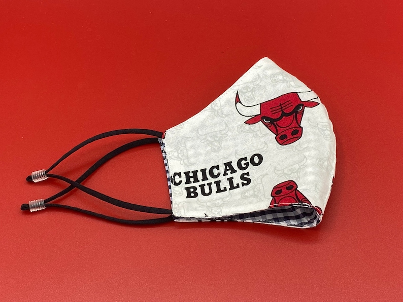 Puede incluir: M&aacute;scara facial blanca con un patr&oacute;n a cuadros blanco y negro en el interior. El exterior de la m&aacute;scara presenta un fondo blanco con un logotipo rojo de los Chicago Bulls y el texto "CHICAGO BULLS". La m&aacute;scara tiene lazos el&aacute;sticos negros para las orejas.