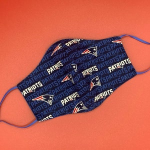 Puede incluir: Una mascarilla facial azul con un patr&oacute;n repetido de la palabra "Patriots" y un casco de f&uacute;tbol estilizado. La mascarilla tiene cintas el&aacute;sticas azules.