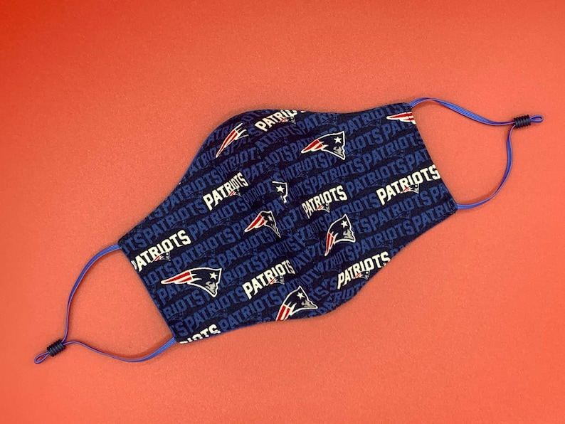 Puede incluir: Una mascarilla facial azul con un patr&oacute;n repetido de la palabra "Patriots" y un casco de f&uacute;tbol estilizado. La mascarilla tiene cintas el&aacute;sticas azules.