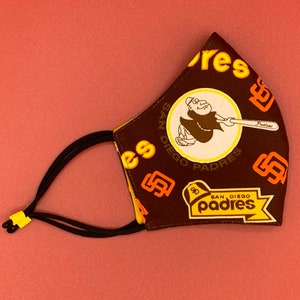 Puede incluir: Máscara facial de tela marrón con un logotipo de los Padres de San Diego amarillo y naranja y el texto "San Diego Padres".