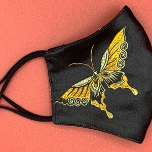 Pode incluir: Máscara facial preta com um design de borboleta bordada em ouro. A máscara tem tiras elásticas pretas com ajustadores de metal prateados.