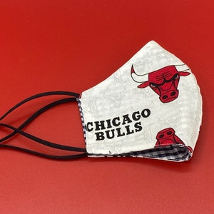 Puede incluir: M&aacute;scara facial blanca con un patr&oacute;n a cuadros blanco y negro en el interior. El exterior de la m&aacute;scara presenta un fondo blanco con un logotipo rojo de los Chicago Bulls y el texto "CHICAGO BULLS". La m&aacute;scara tiene lazos el&aacute;sticos negros para las orejas.