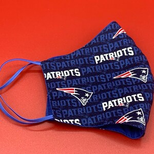 Puede incluir: Una mascarilla facial azul con un patr&oacute;n repetido del logotipo de los New England Patriots y la palabra "Patriots" en blanco. La mascarilla tiene lazos el&aacute;sticos para las orejas de color azul.