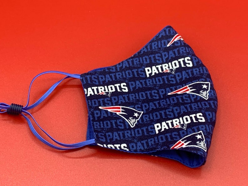 Puede incluir: Una mascarilla facial azul con un patr&oacute;n repetido del logotipo de los New England Patriots y la palabra "Patriots" en blanco. La mascarilla tiene lazos el&aacute;sticos para las orejas de color azul.