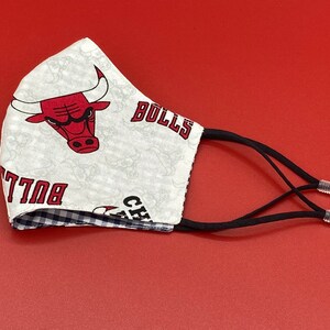 Puede incluir: M&aacute;scara facial blanca con un logotipo rojo de los Chicago Bulls y el texto "Chicago Bulls" impreso en ella. La m&aacute;scara tiene lazos el&aacute;sticos negros para las orejas.