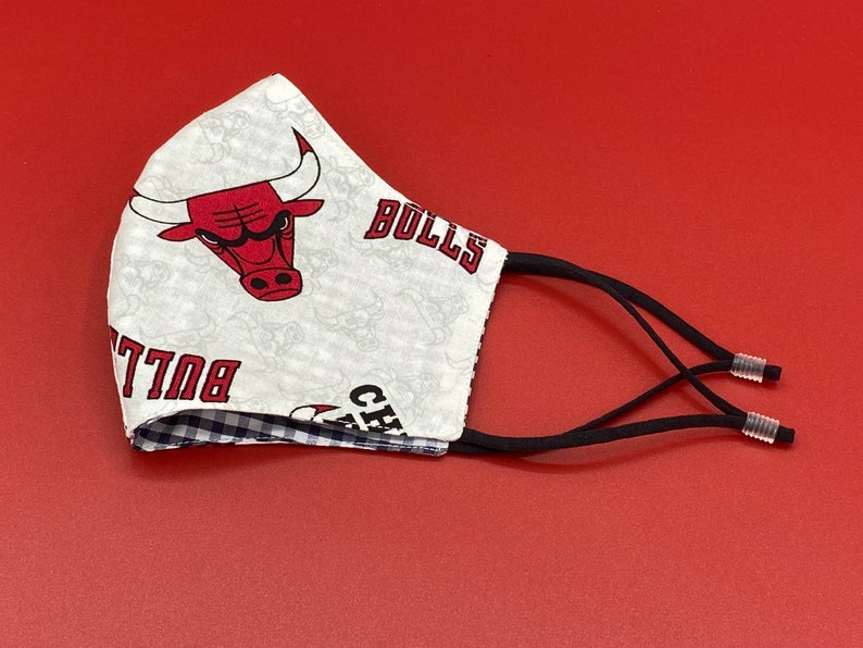 Puede incluir: M&aacute;scara facial blanca con un logotipo rojo de los Chicago Bulls y el texto "Chicago Bulls" impreso en ella. La m&aacute;scara tiene lazos el&aacute;sticos negros para las orejas.