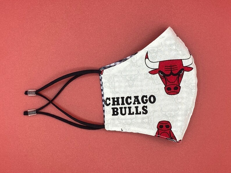 Puede incluir: M&aacute;scara facial blanca con un patr&oacute;n de cuadros blanco y negro y un logotipo rojo de los Chicago Bulls con el texto "CHICAGO BULLS". La m&aacute;scara tiene lazos el&aacute;sticos negros para las orejas.