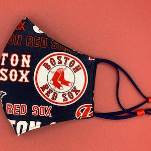 May include: A blue face mask with a white and red Boston Red Sox logo and repeated text "Red Sox" and "Boston". The mask has navy blue ties with red plastic adjusters.