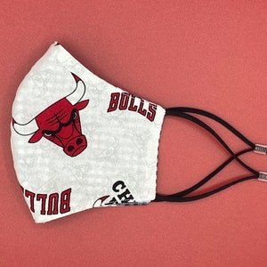 Puede incluir: M&aacute;scara facial blanca con un logotipo rojo de los Chicago Bulls y la palabra "Bulls" impresa en ella. La m&aacute;scara tiene lazos el&aacute;sticos negros para las orejas.
