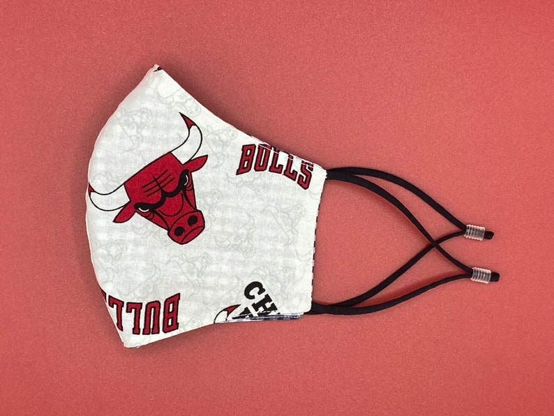 Puede incluir: M&aacute;scara facial blanca con un logotipo rojo de los Chicago Bulls y la palabra "Bulls" impresa en ella. La m&aacute;scara tiene lazos el&aacute;sticos negros para las orejas.