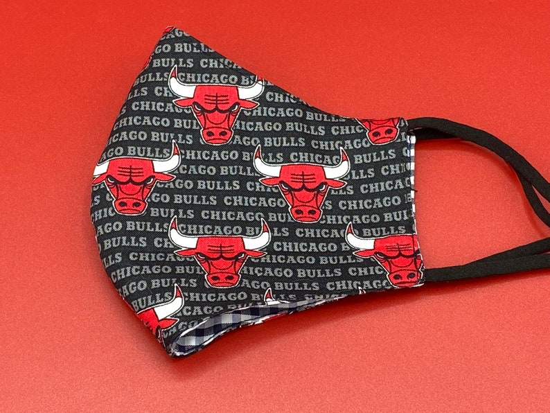May include: Black face mask with a repeating pattern of the Chicago Bulls logo. The logo features a red bull head with black horns and a white outline. The text "Chicago Bulls" is repeated in a gray font on the black background.