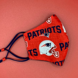 Puede incluir: M&aacute;scara facial roja con un logotipo de los New England Patriots en blanco y azul y la palabra "Patriots" impresa en ella. La m&aacute;scara tiene lazos azul marino.