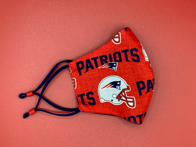 Puede incluir: M&aacute;scara facial roja con un logotipo de los New England Patriots en blanco y azul y la palabra "Patriots" impresa en ella. La m&aacute;scara tiene lazos azul marino.