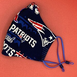 Puede incluir: Una mascarilla facial azul con un logotipo "Patriots" blanco y estrellas rojas, blancas y azules. La mascarilla tiene lazos el&aacute;sticos azules para las orejas.