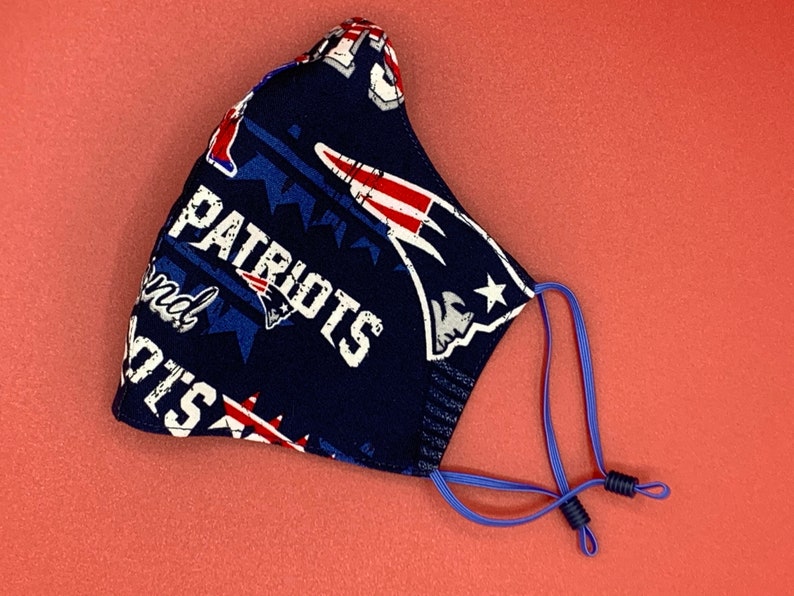 Puede incluir: Una mascarilla facial azul con un logotipo "Patriots" blanco y estrellas rojas, blancas y azules. La mascarilla tiene lazos el&aacute;sticos azules para las orejas.