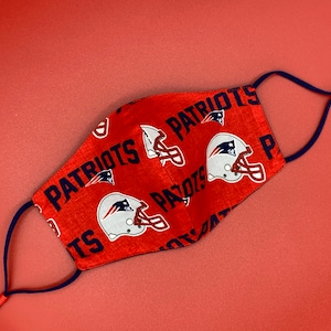 Puede incluir: M&aacute;scara facial roja con un patr&oacute;n repetido del logotipo de los New England Patriots y la palabra "Patriots". La m&aacute;scara tiene lazos ajustables para las orejas de color azul marino.