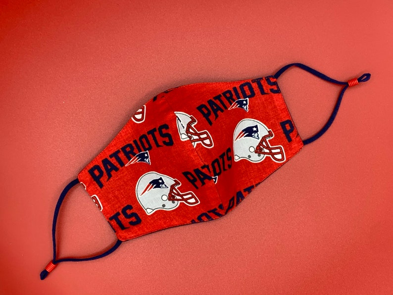 Puede incluir: M&aacute;scara facial roja con un patr&oacute;n repetido del logotipo de los New England Patriots y la palabra "Patriots". La m&aacute;scara tiene lazos ajustables para las orejas de color azul marino.