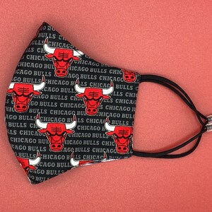 Puede incluir: M&aacute;scara facial negra con un patr&oacute;n repetido del logotipo de los Chicago Bulls y las palabras "Chicago Bulls". La m&aacute;scara tiene correas el&aacute;sticas negras con ajustadores plateados.