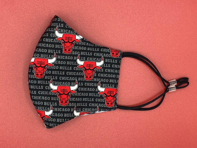 Puede incluir: M&aacute;scara facial negra con un patr&oacute;n repetido del logotipo de los Chicago Bulls y las palabras "Chicago Bulls". La m&aacute;scara tiene correas el&aacute;sticas negras con ajustadores plateados.