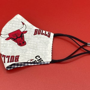 Puede incluir: Una mascarilla facial blanca con un logotipo rojo de los Chicago Bulls y el texto "BULLS" y "CHICAGO" impreso en ella. La mascarilla tiene lazos el&aacute;sticos negros para las orejas con botones ajustables.