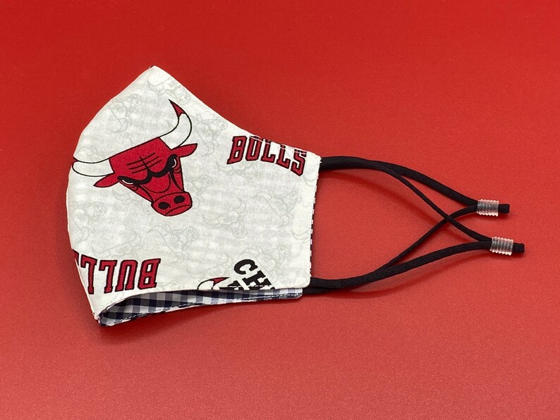 Puede incluir: Una mascarilla facial blanca con un logotipo rojo de los Chicago Bulls y el texto "BULLS" y "CHICAGO" impreso en ella. La mascarilla tiene lazos el&aacute;sticos negros para las orejas con botones ajustables.