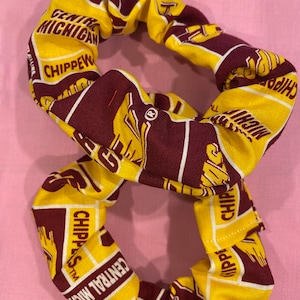 May include: A yellow and maroon fabric scrunchie with the words "Central Michigan Chippewas" printed on it. The scrunchie is in a figure-eight shape, with the fabric folded and sewn to create a gathered effect. The background is a light pink fabric.