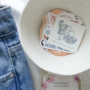 Puede incluir: Un taz&oacute;n blanco lleno de pegatinas coloridas con ilustraciones de flores, mariposas y una ni&ntilde;a regando plantas. Las pegatinas tienen mensajes inspiradores como "Let" y "There is purpose in the wait". El taz&oacute;n est&aacute; sobre una superficie blanca junto a una tarjeta que dice "pegatinas llenas de fe hechas a mano con amor".